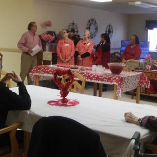 A group of people gathered indoors for an event, featuring a table with decorations and an audience listening to a speaker.