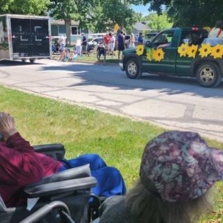 Spectators watch a parade from the sidewalk, where a decorated vehicle with sunflowers passes by.