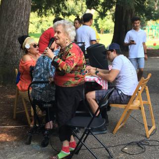 An elderly woman holds a microphone during an outdoor gathering with people seated around her.