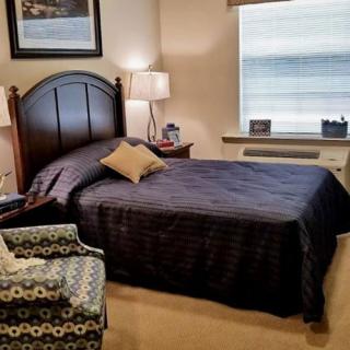 A cozy bedroom with a neatly made bed, a patterned chair, and a window allowing natural light in.