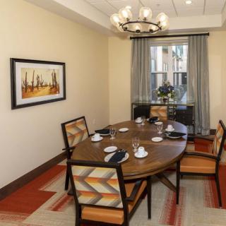 A cozy dining room featuring a wooden table set with black napkins and cups, surrounded by cushioned chairs. A chandelier hangs overhead, and a piece of artwork decorates the wall.