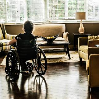 An elderly person in a wheelchair gazes thoughtfully in a cozy living room filled with natural light.