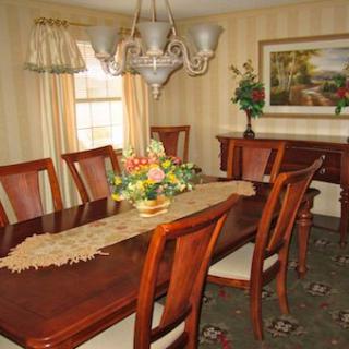 A traditional dining room with a wooden table, chairs, and chandelier, decorated with flowers and curtains.