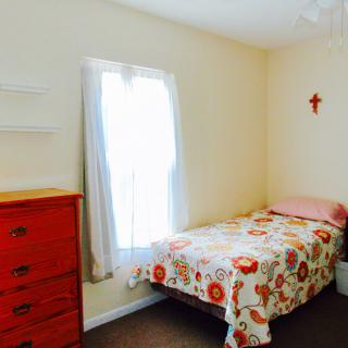 A cozy bedroom with a single bed featuring floral bedding, a dresser, and a window allowing natural light to enter.