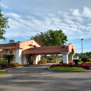 A building with a covered entrance and landscaped surroundings, featuring colorful flowers and trees under a cloudy sky.