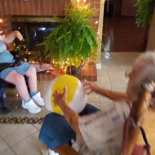 Two people playing with a beach ball indoors, one seated in a wheelchair near a fireplace.