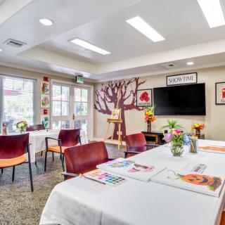 A well-lit room with tables set up for art activities, featuring colorful chairs, decorations, and a television on the wall.