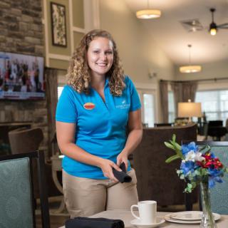 A smiling woman in a uniform setting a table in a restaurant with floral decoration.