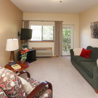 A cozy living room with a sofa, armchair, and television, featuring warm lighting and a window overlooking greenery.