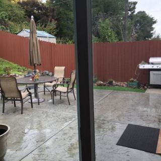 A backyard patio with a table, chairs, umbrella, and a grill against a wooden fence.