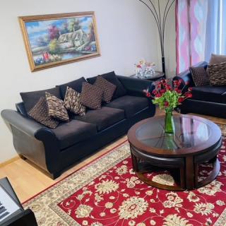 A cozy living room featuring a dark sofa set, a round coffee table with a vase of flowers, and a patterned red rug.