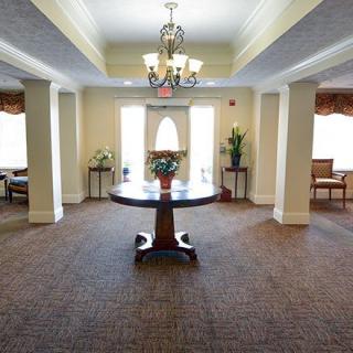 A well-lit interior space featuring a round table with floral decoration, surrounded by windows and elegant furnishings.