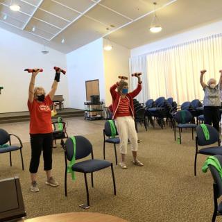 A group of people participating in an indoor fitness class, using dumbbells and chairs for their exercise routine.