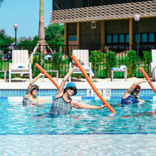 A group of people participating in a water aerobics class in an outdoor pool, using foam noodles for exercise.