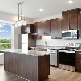 A modern kitchen with a central island, stainless steel appliances, wooden cabinets, and large windows providing natural light.