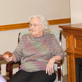 An elderly woman sits comfortably in an armchair next to a fireplace, smiling warmly.