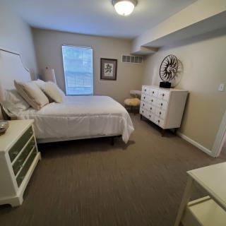 A cozy bedroom with neutral tones, featuring a bed with multiple pillows, a dresser, and decorative items.