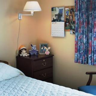 A cozy bedroom with a neatly made bed, lamp, and a nightstand decorated with stuffed toys and family photos.