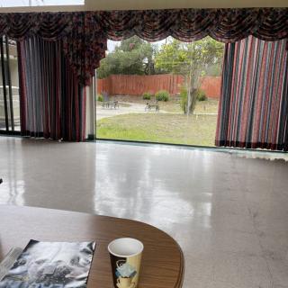 A spacious room with large windows overlooking a garden, featuring a table with a paper cup and magazine.