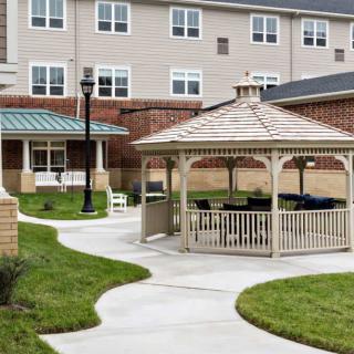 A community outdoor area featuring a gazebo and pathways surrounded by green grass, adjacent to a building.