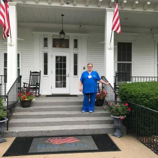 A person in a nurse's uniform stands on the steps of a building with American flags.