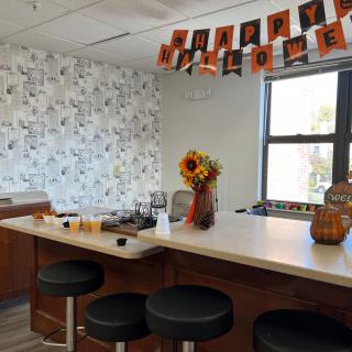 A kitchen area decorated for Halloween with a 'Happy Halloween' banner, sunflowers in a vase, and pumpkins on the counter.