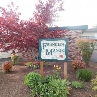 A sign reading 'Franklin Manor' stands amidst autumnal landscaping with a red-leaved tree and surrounding bushes in front of a building.