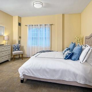 A cozy bedroom with soft yellow walls, featuring a neatly made bed adorned with blue pillows, a dresser, and a window with sheer curtains.