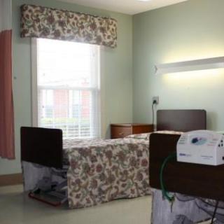 A view of a hospital patient room featuring a bed, window with floral curtains, and medical equipment.