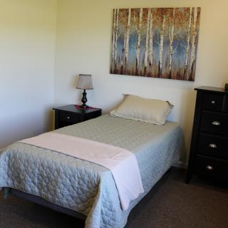 A minimalist bedroom featuring a single bed with light bedding, a nightstand with a lamp, and framed artwork on the wall.