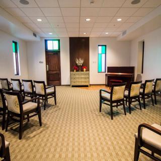 A small chapel with arranged seating and a piano, featuring carpeted flooring and windows with colored glass.