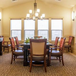 A well-lit dining room with a large table surrounded by chairs, featuring multiple windows and a central chandelier.
