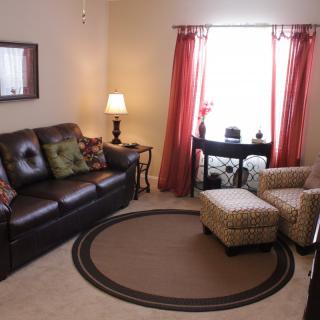 A cozy living room with a brown leather sofa, a patterned armchair and ottoman, warm lighting from a lamp, and red curtains framing the window.