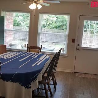A dining area with a table covered in a blue tablecloth, surrounded by chairs, and lit by natural light from large windows.