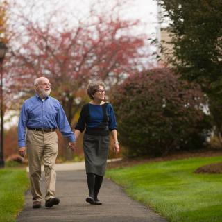 An elderly couple walks hand in hand through a park on an autumn day.