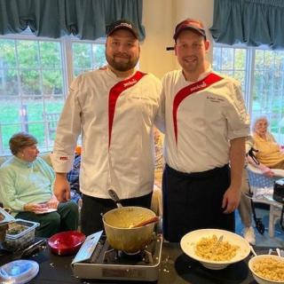 Two chefs in uniform standing by a cooking station with a pasta dish prepared.