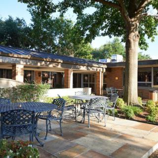 A peaceful outdoor patio area with metal furniture, surrounded by greenery and a building in the background.