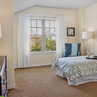 A cozy bedroom with a large window, featuring a bed with patterned bedding and a comfortable chair.