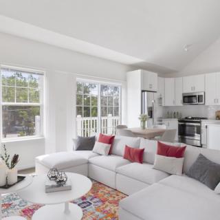A modern open-concept living room with a white sectional sofa, colorful throw pillows, and a bright kitchen area.
