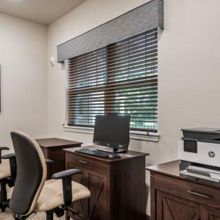 A small office workspace featuring desks with a computer and a printer, two chairs, and a window with blinds.