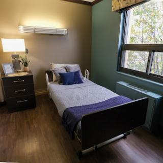 A neatly arranged hospital room featuring a single bed, bedside lamp, and natural light from the window.
