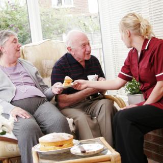 A caregiver interacting with an elderly couple during tea time.