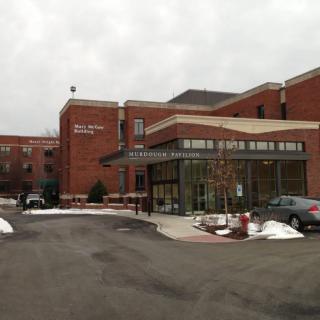 A hospital building with a brick exterior, featuring an entrance and adjacent parking lot with some snow on the ground.
