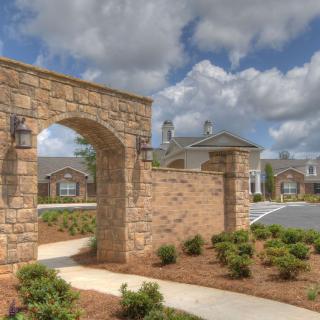 A picturesque stone archway leads to a residential community, surrounded by neatly maintained landscaping under a cloudy sky.