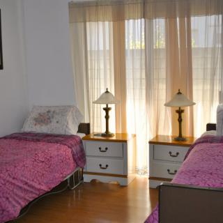 A cozy bedroom with two twin beds, each adorned with pink bedspreads, flanking a central nightstand under sheer curtains letting in daylight.
