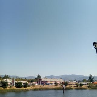 A serene view of a lakeside landscape with buildings and mountains in the background under a clear blue sky.