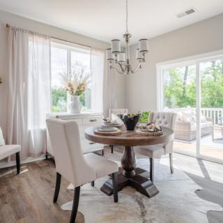 A bright and elegantly decorated dining room with a round table, upholstered chairs, and a chandelier, illuminated by natural light from large windows.