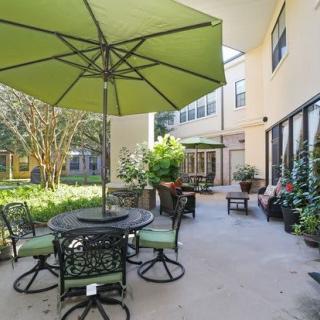 A well-furnished outdoor patio area with a table, chairs, and a large green umbrella surrounded by greenery.