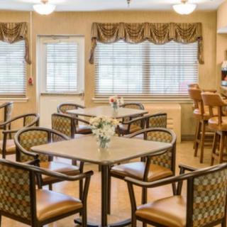 A cozy dining area with multiple tables and chairs, featuring large windows with blinds and decorative lighting.
