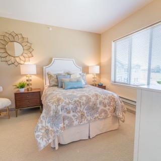 A cozy and well-decorated bedroom with neutral tones, featuring a bed with patterned bedding, two bedside lamps, and a large window letting in natural light.